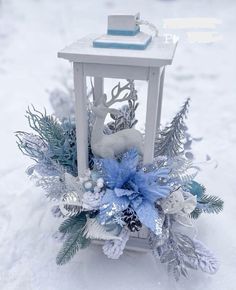 a small white lantern in the snow with blue flowers and pineconis on it