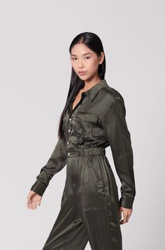 This utility jumpsuit is a chic and practical garment that combines modern style with functional design. The jumpsuit is rendered in a deep olive green colour, providing a versatile and earthy aesthetic. It features a collared neckline and a button-down front with off white buttons. The waistline is cinched with an elastic band, creating a defined silhouette that flatters the figure while maintaining comfort. The trouser, which form the lower part of the jumpsuit, are tailored with an adjustable tapered cuff that can been worn open or closed depending on your shoe choice. Designed in Hong Kong Made ethically in China Fits true to size Sand-Washed Mulberry Silk Small Batch Sustainable Finished with Internal French Seams International Certification: OEKO-TEX Standard 100  Care Guide Dry Clea Green Utility Jumpsuits And Rompers For Work, Green Utility Style Jumpsuit For Work, Green Utility Jumpsuit For Workwear, Green Utility Jumpsuit With Long Sleeves, Green Long Sleeve Utility Jumpsuit, Fitted Utility Jumpsuit With Button-up Design, Deep Olive Green, Khaki Military Long Sleeve Jumpsuit, Fitted Button-up Utility Jumpsuit/romper