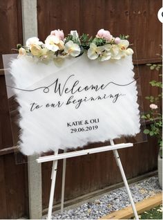 a welcome sign with flowers on it in front of a wooden fence and some gravel