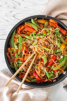 a pan filled with noodles and vegetables on top of a white wooden table next to chopsticks