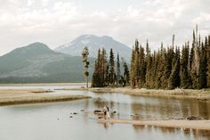 two people are standing on the shore of a lake in front of some trees and mountains