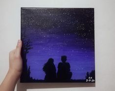 a hand holding up a painting with two people looking at the stars