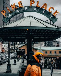 a man in an orange jacket standing under a green and white sign that says pike place