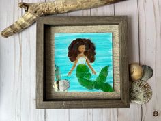 a painting of a woman with long hair and green leaves in a frame next to seashells