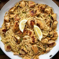 a white plate topped with pasta and chicken next to lemon wedges on top of a wooden table