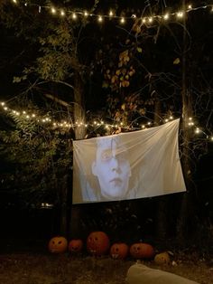 an outdoor movie screen with pumpkins in front of it and lights strung from the trees