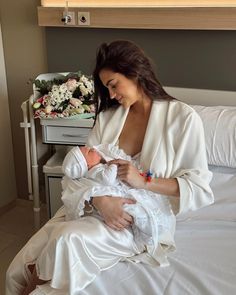 a woman holding a baby in her arms while sitting on a bed next to flowers