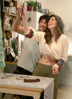 a man and woman taking a selfie in the kitchen with chocolate cake on the table