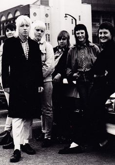 a group of people standing next to each other near a car on a street with buildings in the background