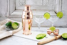 a copper cocktail shaker with limes on the side next to it and two glasses