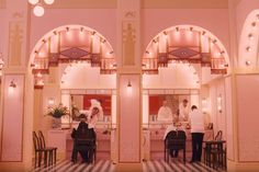 two people standing in front of a pink building with arches and lights on the ceiling