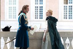 two people dressed in period clothing talking to each other on a balcony overlooking a building