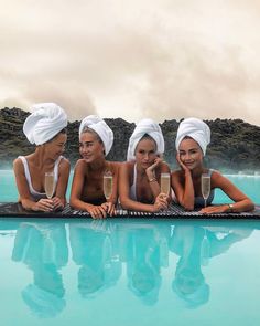 four beautiful women sitting on the edge of a swimming pool with champagne in their hands