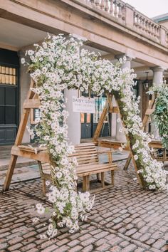 a wooden bench sitting on top of a brick floor next to a white flower covered arch