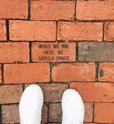 a person standing in front of a brick wall with the words while we are here, we should dance