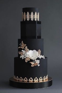 a three tiered black cake with white flowers and leaves on the top, against a gray background