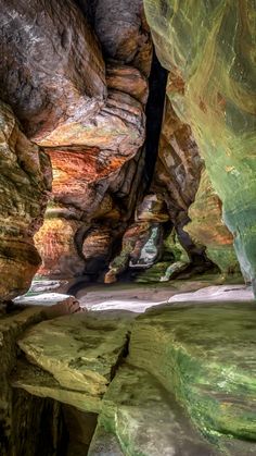 the inside of a cave with green and brown rocks, water and sand on the ground