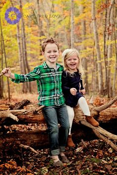 two children sitting on a fallen tree in the woods with fall leaves and foliage around them