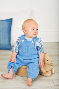 a baby sitting on the floor next to a teddy bear and wearing blue overalls