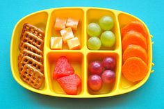 a yellow tray with fruit, crackers and pretzels in it on a blue surface