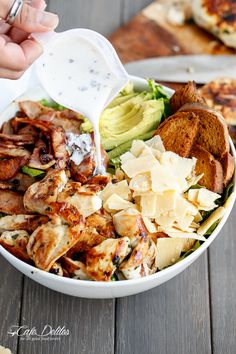 someone pouring ranch dressing into a bowl of chicken, avocado and toasted bread