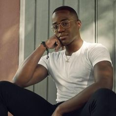 a man sitting on the ground with his hand under his chin, wearing glasses and a white t - shirt