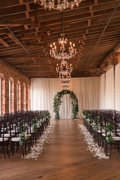 an indoor wedding venue with rows of chairs and chandeliers, decorated with greenery