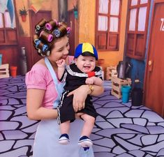 a woman holding a baby in front of a fake room with toys on the floor