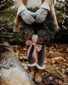 a woman holding a wreath with pine cones and bows on it in front of some trees