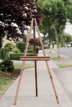 a wooden easel sitting on the side of a sidewalk next to a tree and grass