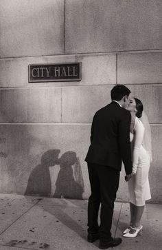 a black and white photo of a couple kissing in front of the city hall sign