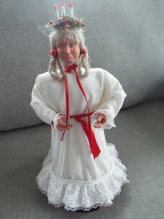 a doll dressed in white and wearing a red ribbon around her neck, standing on a gray couch