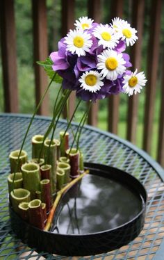 purple and white flowers are in a black tray on a table with bamboo sticks as vases