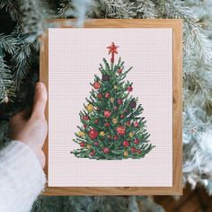 a cross stitch christmas tree is displayed on a wooden frame in front of a pine tree