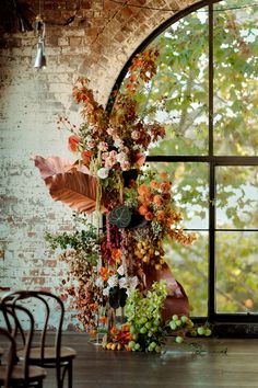 an arrangement of flowers and foliage in front of a large window on a brick wall