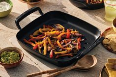 a pan filled with assorted vegetables on top of a table next to bowls and spoons