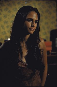 a woman with long dark hair wearing a brown dress and looking at the camera while standing in front of a wall