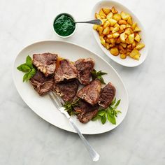 two plates with meat and potatoes on them next to a bowl of green dipping sauce