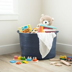 a teddy bear sitting in a blue basket filled with books and toys next to a window