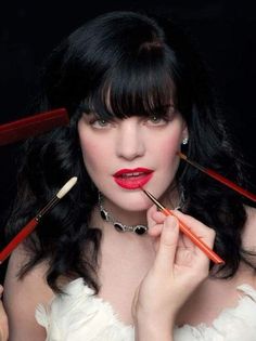 a woman with long black hair and red lipstick holding two brushes to her face, while she is looking at the camera