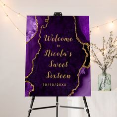 a purple and gold welcome sign on a easel next to a vase with flowers