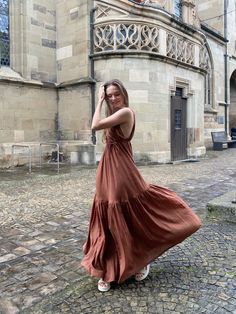 a woman in a long brown dress standing on a cobblestone street next to an old building