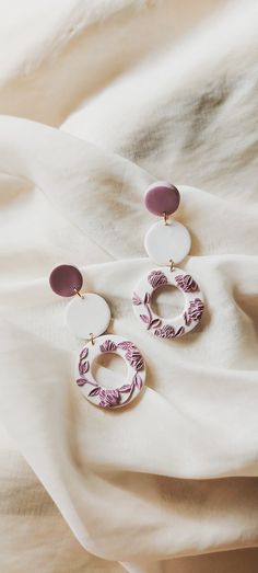 a pair of earrings on top of a white sheet with purple and white designs hanging from it