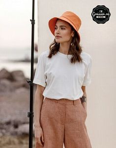 a woman standing next to a white wall wearing an orange hat and wide legged pants