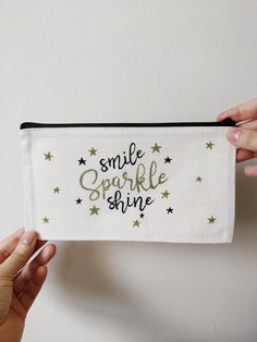 "Elevate your daily essentials with this unique \"Smile Sparkle Shine\" pouch, meticulously handwritten with waterproof gold and black pens on soft and durable canvas. The sturdy metal zipper ensures smooth closure, making it perfect for everyday use and an excellent choice for gifting something truly special. This practical and durable canvas pouch serves a multitude of purposes. It can house your daily makeup essentials, making it an ideal cosmetic pouch for travel and school. Additionally, it's perfect for storing jewelry, acting as a pencil case, wallet, or compact toiletry bag. ✨Product Specifications: ・Material: Canvas and Metal Zipper ・Size: Approx. 21 x 12 cm (8.4 x 4.8 inches) ・Color: Beige background with a sleek black zipper With just one available, don't miss the opportunity to Cricut Pouch Bag Ideas, Smile Sparkle Shine, Projet Cricut, Pouch Ideas, Apparel Business, Black Pens, Bag Painting, Custom Makeup Bags, Vinyl Printing