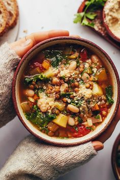 a person holding a bowl of soup with beans and spinach in it, next to some bread