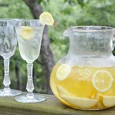 three glasses and a pitcher with lemons in them sitting on a table outside next to trees