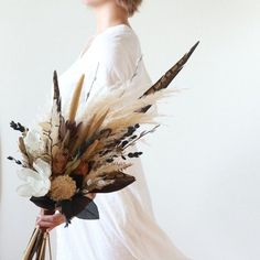 a woman holding a bouquet of dried flowers and feathers in her left hand, standing against a white wall