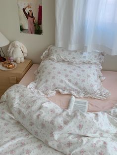 an unmade bed with pink and white flowers on it, next to a window