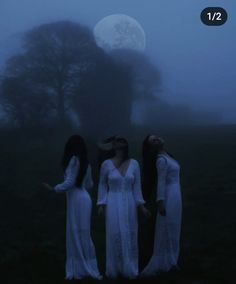 three women in white dresses are standing on the grass at night, with trees behind them